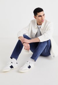 Chaqueta blanca, camiseta gris y jeans azules llevados por una figura sentada. Se ven zapatillas blancas con acentos en azul marino y suelas texturizadas.