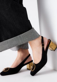 Zapatos de charol negros tipo slingback con punta redonda, que cuentan con un emblema decorativo dorado y un tacón cuadrado con acentos dorados y tachuelas.