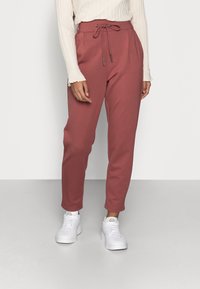 Person som bär högmidjade, dammrosafärgade tapered byxor med dragsko i midjan, krämfärgad ribbad tröja och vita sneakers som står på en grå golv.