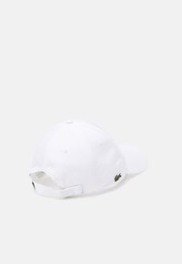 Hvit bomulls baseballcap med justerbar stropp, med en liten grønn krokodille-logo på siden og ventilasjonsøyler på toppen.