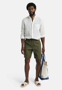 Weißes Leinenhemd, grüne Shorts mit Kordelzug und beiges Rutschesandalen. Hält eine zweifarbige Tasche mit blauen Griffen.