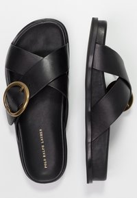 Černé kožené sandály na klínu s kříženými popruhy a velkým bronzovým kroužkem jako akcentem. Plná podrážka s minimalistickým designem a brandingem.