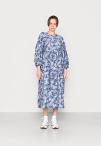 Vestido azul com padrão floral, mangas bufantes longas, cintura franzida e saia fluída, combinado com ténis brancos de altura até ao tornozelo.