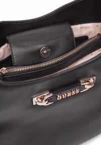 Sac à main en cuir noir avec un compartiment zippé et un rabat repliable. Quincaillerie en ton doré et détail de marque en relief.