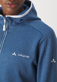 Modra zip-up kapuca, izdelana iz teksturirane tkanine, z belim kontrastnim obrobom kapuce, zadrgo in logotipom "VAUDE" na prsih ter na rokavu.