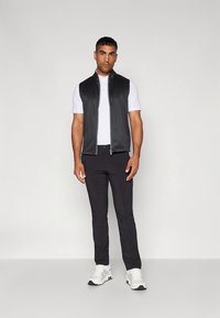 Črni brezrokavnik čez belo polo majico, kombiniran s črnimi hlačami in belimi supergami. Gladek tekstura z modernim dizajnom.