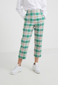 Osoba w białych trampkach, skróconych zielono-pomarańczowych spodniach w kratę oraz białej koszuli z długim rękawem z zapięciem na guziki, z rękami w kieszeniach.