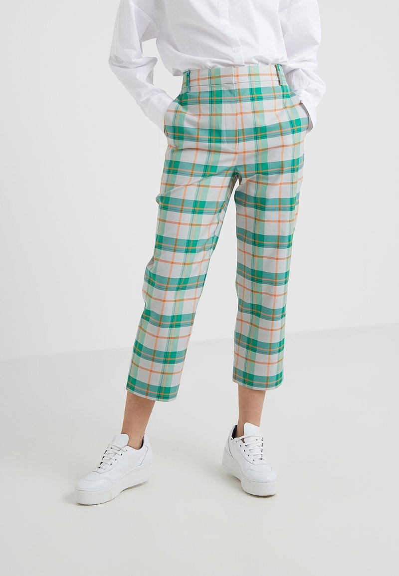 Osoba w białych trampkach, skróconych zielono-pomarańczowych spodniach w kratę oraz białej koszuli z długim rękawem z zapięciem na guziki, z rękami w kieszeniach.