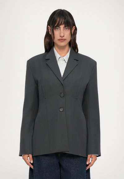 Femme portant un blazer oversize gris foncé à deux boutons sur une chemise gris clair et un jean foncé, se tenant devant un fond blanc uni.