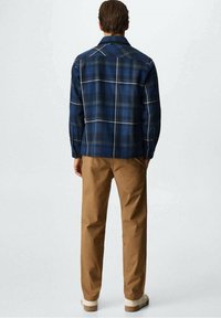 Camicia a quadri blu navy con cuciture chiare, dotata di colletto e maniche lunghe, abbinata a pantaloni khaki e calzature beige.