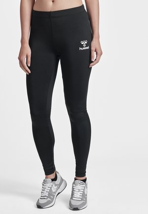 Vrouw draagt zwarte Hummel legging en grijs-witte Hummel Racer sneakers, staand tegen een effen lichte achtergrond.