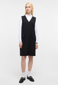 Zwarte mouwloze jurk met een V-hals, voorzien van twee zijzakken en een gladde textuur; gecombineerd met een witte overhemd met kraag en loafers.
