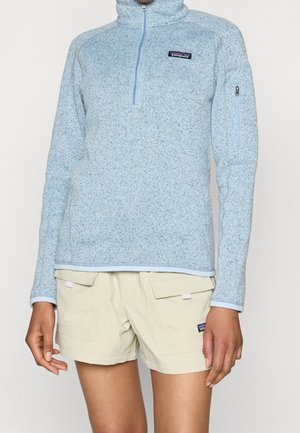 Lyseblå fleece med kvart-lyn-lås og en lille lynlåslomme på ærmet, båret sammen med beige cargoshorts med klaplommer.