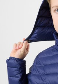 Laivastonsininen toppatakki irrotettavalla hupulla, jossa on tikattu pinta ja napin kiinnitys kaulassa. Läheltä kuvattu käsi, joka pitää huppua kiinni.