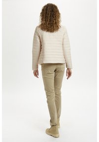 Jasnoróżowa pikowana kurtka o pudełkowym kroju, zestawiona z beżowymi spodniami i beżowymi botkami. Widok od tyłu podkreśla fakturę tkaniny i detale szwów.