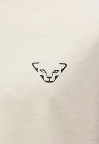 Μαύρο μινιμαλιστικό λογότυπο προσώπου τσιτάχ στο κέντρο σε φόντο κρεμ υφάσματος.