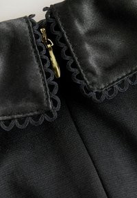 Melnas audums ar viļņota apkakles detaļu un zelta rāvējslēdzēju. Faktūra ir gluda ar tēlaini spīdumu.