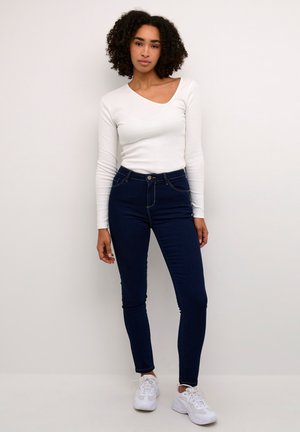 White long-sleeve top paired with dark blue skinny jeans. The jeans feature a high waist and subtle stitching details. Footwear is white sneakers.