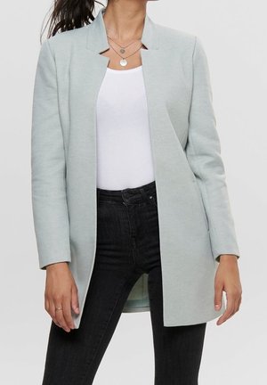 Femme portant un blazer gris clair ouvert sur un top blanc et un jean noir taille haute, avec des colliers argentés superposés visibles.