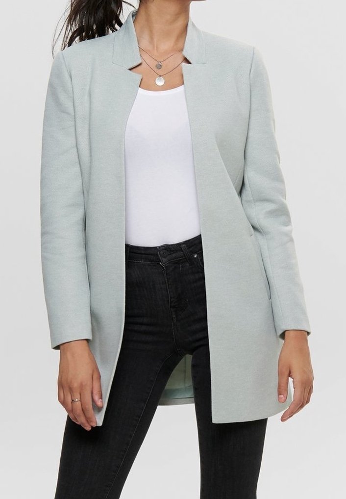 Femme portant un blazer gris clair ouvert sur un top blanc et un jean noir taille haute, avec des colliers argentés superposés visibles.