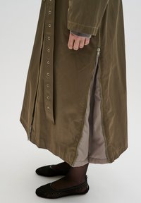 Cappotto lungo verde oliva con cerniera verticale, bottoni automatici e interno di colore chiaro a contrasto. Indossato con scarpe mesh nere.