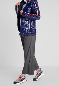 Giubbotto imbottito blu con finitura lucida, strisce laterali bianche e rosse, cappuccio, chiusura con zip, abbinato a pantaloni larghi grigi e sneakers colorate.