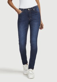 Dunkelblaue Skinny-Jeans mit einer verblassten Textur, fünf Taschen und kontrastierenden Nahtdetails. Kombiniert mit weißen Sneakers.