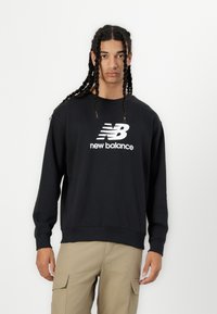 Črna bombažna majica s dolgimi rokavi, z rebrastimi manšetami in okroglim izrezom. Ima velik bel logotip "New Balance" na prsih.
