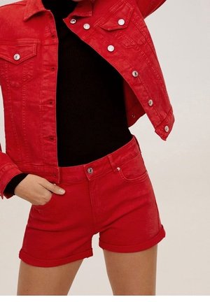 Zbliżenie osoby ubranej w czerwoną kurtkę z denimu i pasujące czerwone szorty z denimu z srebrnymi guzikami, noszone na czarnym topie.
