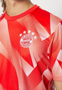 Camisa deportiva roja con un patrón geométrico y acentos blancos, cuello redondo y el logo del FC Bayern München con cuatro estrellas encima.