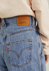 Valaise sinivärvi teksad tekstuuriga viimistlusega, millel on pruun nahkplekk vööl ja iseloomulik tagasi tasku õmbluse disain.