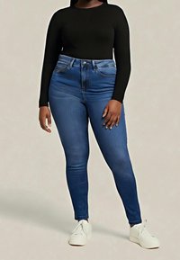 Personne portant un jean skinny bleu foncé, un haut à manches longues noir et des baskets blanches, debout devant un fond beige uni.