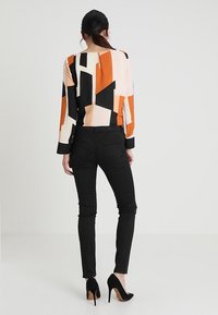 Femme aux cheveux noirs attachés en queue de cheval basse, portant un haut à manches longues géométrique orange, noir et beige, un pantalon noir ajusté et des talons noirs.