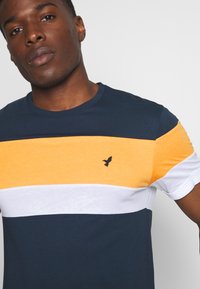 Navy blue, yellow, and white striped short-sleeve t-shirt. Features round neckline and embroidered bird logo on the chest. Casual fit.