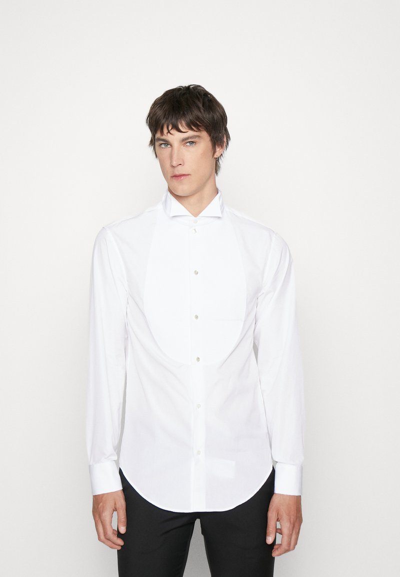 Chemise blanche avec col en aile, manches longues et ourlet arrondi. Comprend un plastron à l'avant et une fermeture à boutons. Texture de tissu lisse.