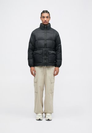 Giacca a vento nera con colletto alto, zip frontale e due tasche anteriori. Abbinata a pantaloni cargo beige e sneaker bianche.