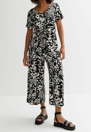 Vrouw draagt een zwart-wit abstracte print korte mouw jumpsuit en zwarte platformsandalen, staand tegen een effen achtergrond.