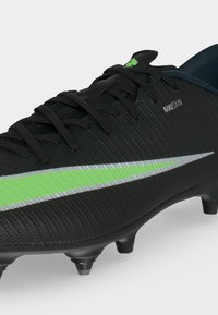 Μαύρο ποδοσφαιρικό παπούτσι Nike με πράσινο και ασημί σήμα, μαύρα κορδόνια και σόλα με πέλματα για παιχνίδι σε τεχνητό χλοοτάπητα ή γρασίδι.