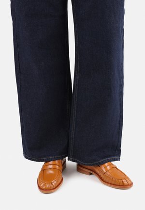 Tan leather loafers with a textured, pleated design, paired with dark blue wide-leg jeans featuring contrasting stitching.