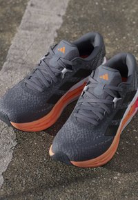 Paire de chaussures de course grises et orange avec des bandes noires sur le trottoir, dotées de collerettes rembourrées et de semelles intermédiaires amorties.
