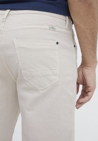 Svijetlosmeđi traperice slim fit kroja, s dva stražnja džepa i tamnim gumbima. Tkanina izgleda mekano s minimalnom teksturom.
