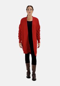 Roter Strickcardigan mit gerippter Textur, langen Ärmeln mit Knopfdesign, offener Front, über einem schwarzen Oberteil und Leggings getragen, kombiniert mit braunen Stiefeln.