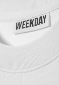 Hvid bomulds T-shirt krave med ribbet tekstur; mærket viser "WEEKDAY" med fede sorte bogstaver. Glat stof, enkelt design.