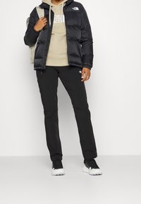 Svart puffjacka med en figursydd silhuett, ljusbeige hoodie med logotyp, svarta byxor och svarta sportskor med vita detaljer.