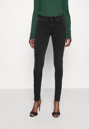 Frau trägt schwarze Skinny-Jeans, grünes langärmeliges Oberteil und schwarze High-Heel Sandalen mit offenen Zehen, steht vor einem einfarbigen Hintergrund.