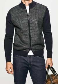 Marineblauer und grauer Sweatshirt mit Kragen und Reißverschluss, mit strukturiertem Karomuster auf der Vorderseite, gestrickten Ärmeln und geripptem Saum, kombiniert mit dunklen Jeans.