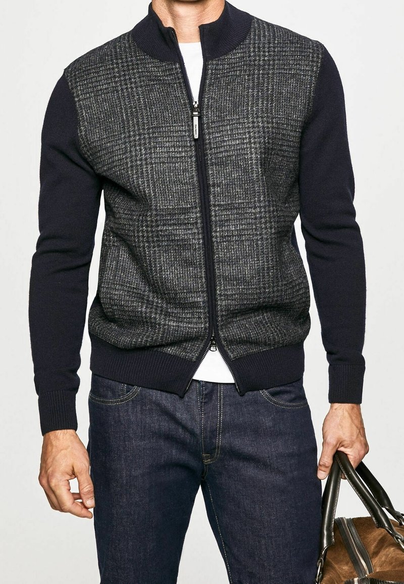 Marineblauer und grauer Sweatshirt mit Kragen und Reißverschluss, mit strukturiertem Karomuster auf der Vorderseite, gestrickten Ärmeln und geripptem Saum, kombiniert mit dunklen Jeans.