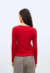 Γυναίκα με μακριά σγουρά σκούρα μαλλιά, ντυμένη με κόκκινο πλεκτό πουλόβερ και ασπρόμαυρο καρό παντελόνι, απεικονιζόμενη από πίσω.