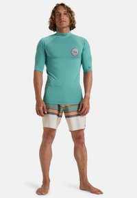 Camisa de surf turquesa de manga corta con un logo y pantalones de baño beige con rayas horizontales de colores. Textura suave, ajuste atlético.