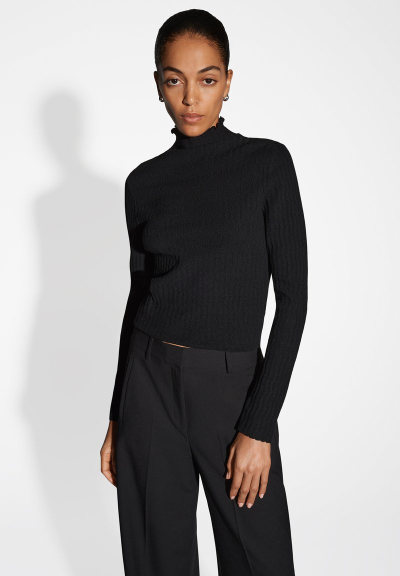 Haut noir à manches longues côtelé avec col montant, présentant une silhouette ajustée et un tissu texturé, associé à un pantalon noir à jambes larges.